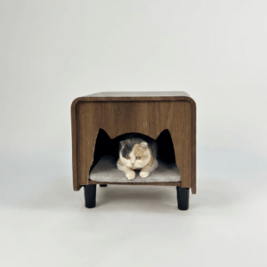 Walnut brown cat nest nightstand with cat-ear entry cutout, black tapered legs, and gray cushion, featuring a calico cat resting inside on a light grey background