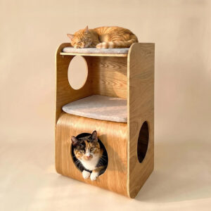 Natural oak multi-level cat tower with oval and circular entry cutouts, three gray cushions, a ginger tabby cat sleeping on top and a calico cat peeking out from the bottom cubby on a beige background